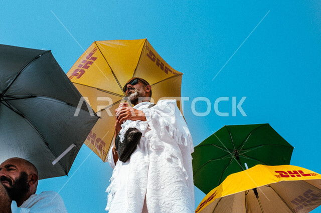 العبادة والتقرب إلى الله بالأعمال الصالحة، شاب خليجي عربي سعودي يرتدي ملابس الاحرام ويحمل بيده مظلة يقف على جبل عرفات، آداء مناسك الحج والعمرة، أماكن ومعالم دينية اسلامية مقدسة في المملكة العربية السعودية