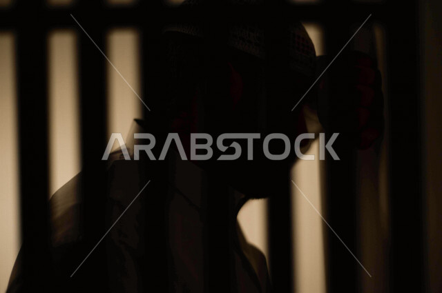 تصوير مقرب لرجل عربي خليجي سعودي ، خلف القضبان الحديدية ، سجين سعودي يضع راسه على الحائط ، الشعور بالندم والكابة ، انتظار الحرية 