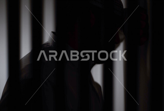 تصوير مقرب لرجل عربي خليجي سعودي ، خلف القضبان الحديدية ، سجين سعودي يضع راسه على الحائط ، الشعور بالندم والكابة ، انتظار الحرية 