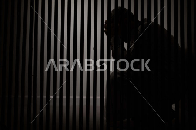رجل عربي خليجي سعودي خلف القضبان الحديدية ، سجين سعودي جالس يضع يده على راسه ، الشعور بالندم والحزن ، ينتظرالحرية 