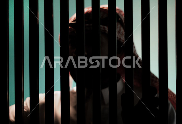 رجل عربي خليجي سعودي داخل السجن ، يمسك القضبان الحديدية ، سجين سعودي ينتظر الحرية ، الشعور بالندم والوحدة 