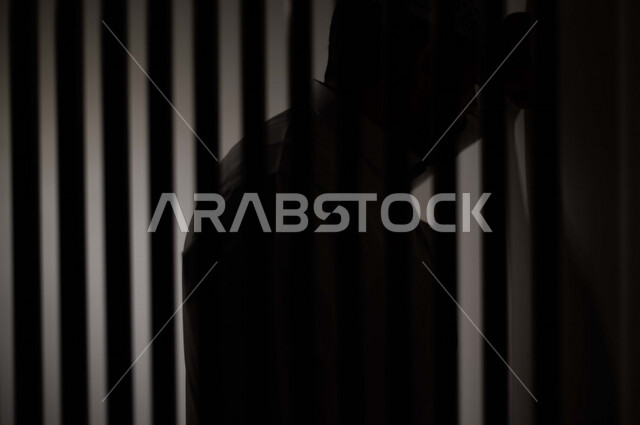 تصوير مقرب لرجل عربي خليجي سعودي ، خلف القضبان الحديدية ، سجين سعودي يضع راسه على الحائط ، الشعور بالندم والكابة ، انتظار الحرية 