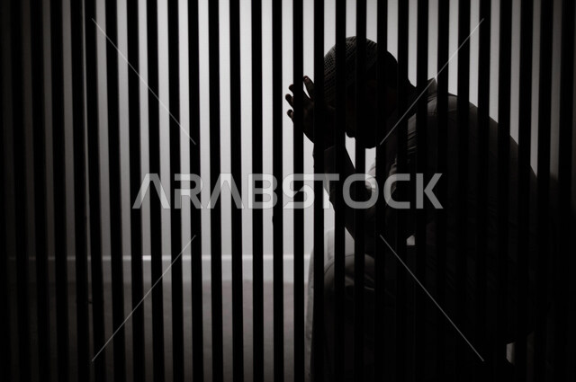 رجل عربي خليجي سعودي خلف القضبان الحديدية ، سجين سعودي جالس يضع يده على راسه ، الشعور بالندم والحزن ، ينتظرالحرية 