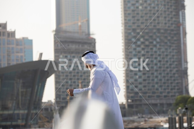 الفن الهندسي المعماري في بناء الأبراج وناطحات السحاب الحديثة, صورة من الجانب لرجل عربي خليجي اماراتي يرتدي الكندورة والغترة يمشي وينظر إلى التطور العمراني في المباني, التأمل والتفكير بالمستقبل, وضع الخطط المستقبلية