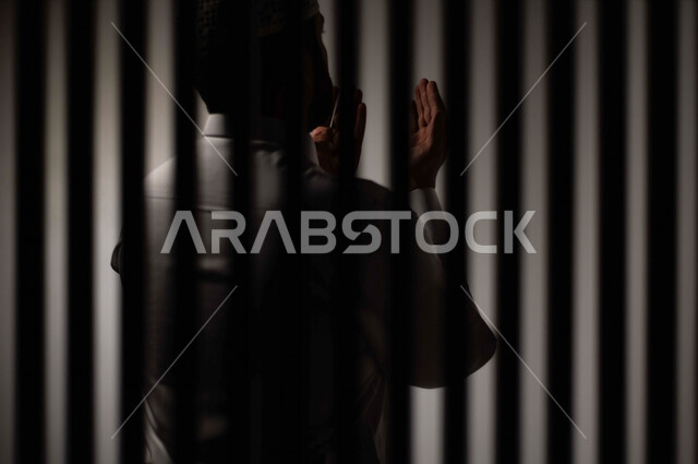 رجل عربي خليجي سعودي خلف قضبان السجن ، يرفع يديه لاعلى يناجي الله، الدعاء والتوبة لله ، صلاة وعبادة