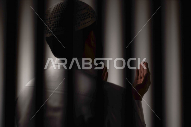 A Saudi Arabian Gulf man behind prison bars, raising his hands up, calling to God, supplication and repentance to God, prayer and worship