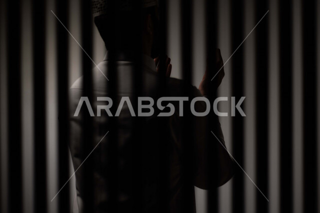 رجل عربي خليجي سعودي خلف قضبان السجن ، يرفع يديه لاعلى يناجي الله، الدعاء والتوبة لله ، صلاة وعبادة