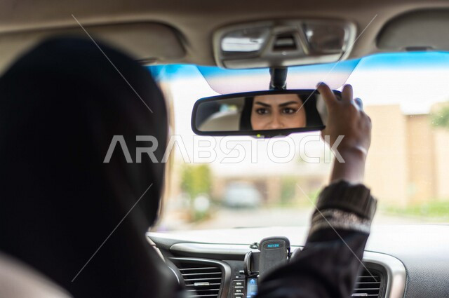 انعكاس على المرآة الامامية للسيارة لامرأة عربية خليجية سعودية محجبة ، تقوم بتعديل المرآة ، امرأة سعودية تقود السيارة ، حرية القيادة للمرأة السعودية ، توصيل الركاب ، راحة وأمان