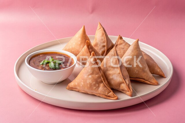 مقبلات رمضانية شهية، فطائر مقلية متنوعة الحشوات، أطباق رئيسية في سفرة إفطار شهر رمضان المبارك، وصفات المأكولات والأطعمة العربية الشرقية السعودية اللذيذة ، طبق من السمبوسك المقرمشة مع صلصة التمر هندي، خلفية وردية