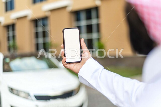 صورة مقربة لرجل عربي خليجي سعودي ، يحمل بيده الهاتف المحمول، يستخدم خدمة النقل من خلال تطبيق التوصيل، تحديد الموقع  ، خدمة توصيل الركاب ، راحة وامان 
