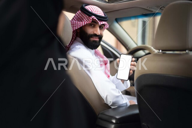 سائق عربي خليجي سعودي يقود السيارة ، يحمل بيده الهاتف المحمول ، امرأة عربية خليجية سعودية محجبة ، تستقل سيارة اجرة ، خدمة النقل من خلال تطبيق التوصيل، تحديد الموقع  ، خدمة توصيل الركاب ، راحة وامان