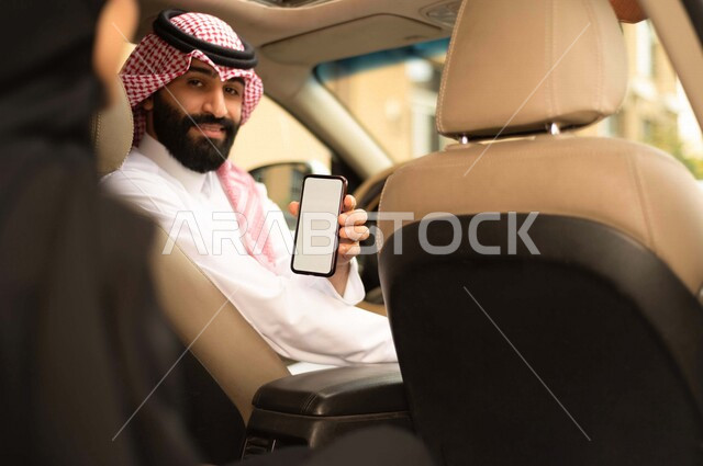 A Saudi Arabian Gulf driver driving the car, holding a mobile phone, a veiled Saudi Arabian Gulf woman, taking a taxi, transportation service through the delivery application, locating, passenger delivery service, comfort and safety