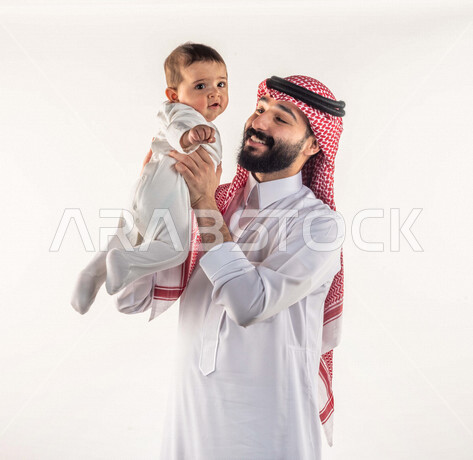 بورتريه لأب سعيد يحمل إبنه الرضيع بيديه لأعلى ، يلعب مع إبنه بإيماءات وجه ويدين مختلفة تدل على السعادة ، رجل عربي سعودي خليجي يرتدي زي سعودي تقليدي مع شماغ وعقال ، خلفية بيضاء ، مفهوم الأبوة والطفولة ، أسرة سعيدة ، تحمل المسؤولية