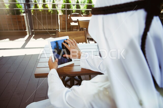 استخدام الاجهزة التقنية الحديثة، ادارة مهام العمل اليومية، صورة مقربة لرجل عربي خليجي سعودي يرتدي الزي التقليدي يجلس في مقهى يحمل بيده جهاز التابلت، مهن ووظائف سعودية