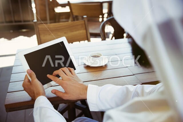 ادارة مهام العمل اليومية، استخدام الاجهزة التقنية الحديثة، صورة مقربة لرجل عربي خليجي سعودي يرتدي الزي التقليدي يجلس في مقهى يحمل بيده جهاز التابلت، مهن ووظائف سعودية