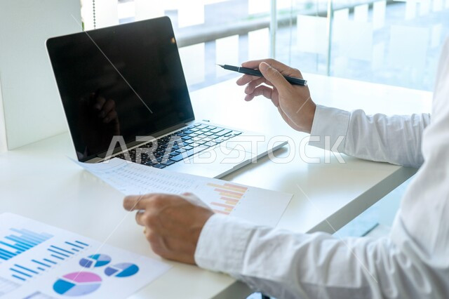 Saudi jobs and professions, using modern technical devices, managing daily work tasks at company headquarters, close-up of a Saudi Arabian Gulf man wearing traditional dress sitting in his office working on a laptop