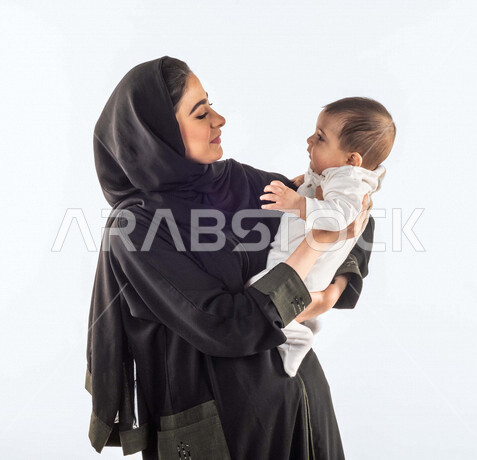 بورتريه لأم تحمل إبنها الرضيع بيديها لأعلى  ، تلعب مع إبنها بإيماءات وجه ويدين مختلفة تدل على السعادة ، إمرأة عربية سعودية خليجية محجبة ، خلفية بيضاء ، مفهوم الأمومة والطفولة ، الأسرة وتحمل المسؤولية