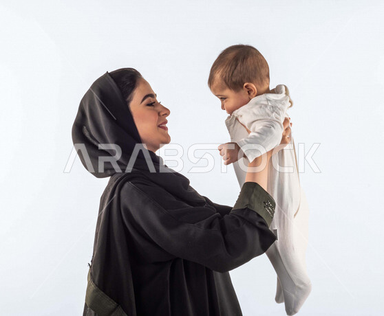 بورتريه لأم تحمل إبنها الرضيع بيديها لأعلى  ، تلعب مع إبنها بإيماءات وجه ويدين مختلفة تدل على السعادة ، إمرأة عربية سعودية خليجية محجبة ، خلفية بيضاء ، مفهوم الأمومة والطفولة ، الأسرة وتحمل المسؤولية