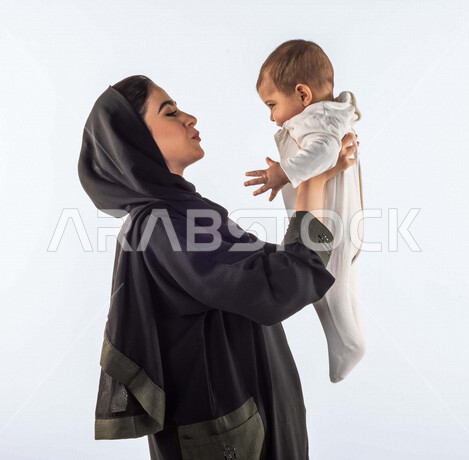 بورتريه لأم تحمل إبنها الرضيع بيديها لأعلى  ، تلعب مع إبنها بإيماءات وجه ويدين مختلفة تدل على السعادة ، إمرأة عربية سعودية خليجية محجبة ، خلفية بيضاء ، مفهوم الأمومة والطفولة ، الأسرة وتحمل المسؤولية