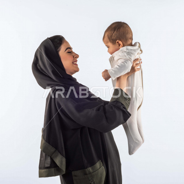 Portrait of a mother holding her infant son with her hands up, playing with her son with different facial and hand gestures indicating happiness, a veiled Saudi Arabian Gulf woman, white background, the concept of motherhood and childhood, family and resp