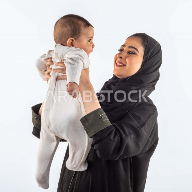بورتريه لأم تحمل إبنها الرضيع بيديها لأعلى  ، تلعب مع إبنها بإيماءات وجه ويدين مختلفة تدل على السعادة ، إمرأة عربية سعودية خليجية محجبة ، خلفية بيضاء ، مفهوم الأمومة والطفولة ، الأسرة وتحمل المسؤولية