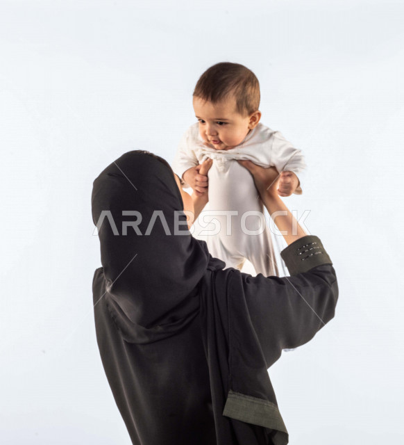 بورتريه لأم تحمل إبنها الرضيع بيديها لأعلى  ، تلعب مع إبنها بإيماءات وجه ويدين مختلفة تدل على السعادة ، إمرأة عربية سعودية خليجية محجبة ، خلفية بيضاء ، مفهوم الأمومة والطفولة ، الأسرة وتحمل المسؤولية