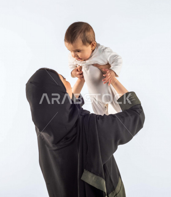 بورتريه لأم تحمل إبنها الرضيع بيديها لأعلى  ، تلعب مع إبنها بإيماءات وجه ويدين مختلفة تدل على السعادة ، إمرأة عربية سعودية خليجية محجبة ، خلفية بيضاء ، مفهوم الأمومة والطفولة ، الأسرة وتحمل المسؤولية