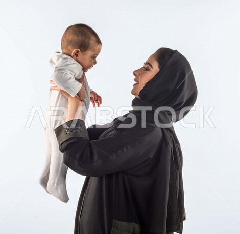 بورتريه لأم تحمل إبنها الرضيع بيديها لأعلى  ، تلعب مع إبنها بإيماءات وجه ويدين مختلفة تدل على السعادة ، إمرأة عربية سعودية خليجية محجبة ، خلفية بيضاء ، مفهوم الأمومة والطفولة ، الأسرة وتحمل المسؤولية