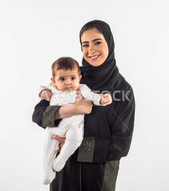 بورتريه لأم تحمل إبنها الرضيع وينظران إلى الكاميرا بإيماءات وجه ويدين مختلفة تدل على السعادة والحب ، إمرأة عربية سعودية خليجية محجبة ، ترتدي عباية سوداء ، خلفية بيضاء ، مفهوم الأمومة والطفولة ، تحمل المسؤولية