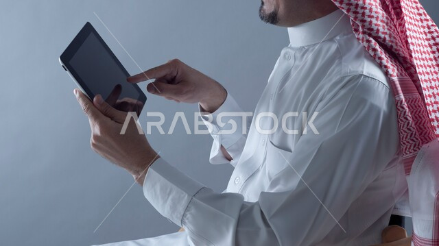 Online shopping using tablet, doing tasks remotely, close-up portrait of a Saudi Arabian Gulf man wearing traditional thobe and shemagh holding a tablet in his hand following social media news, using modern technology and techniques, white background