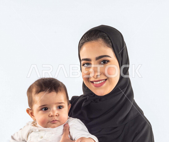 بورتريه لأم تحمل إبنها الرضيع وينظران إلى الكاميرا بإيماءات وجه ويدين مختلفة تدل على السعادة والحب ، إمرأة عربية سعودية خليجية محجبة ، ترتدي عباية سوداء ، خلفية بيضاء ، مفهوم الأمومة والطفولة ، تحمل المسؤولية