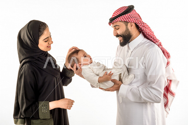 بورتريه لعائلة سعودية خليجية من أب وأم سعيدين بطفلهما، يحمل الأب إبنه الرضيع بين ذراعيه وهو ينظر إليه مع زوجته بإيماءات وجه ويدين مختلفة تدل على الحب والحنان ،خلفية بيضاء ، مفهوم الأسرة السعيدة ، تحمل المسؤولية ، جلسة تصوير لعائلة سعودية صغيرة سعيدة