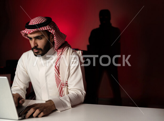 رجل عربي خليجي سعودي يستخدم الحاسوب المحمول ، رجال ملثمين يقفون بالخلف ، ارتكاب اعمال غير قانونية، التجسس السيبراني، هجوم الكتروني ، سرقة بيانات ، الامن السيبراني