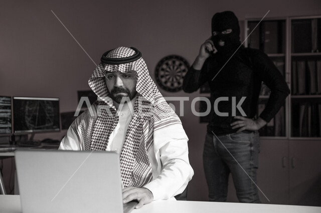 رجل عربي خليجي سعودي يستخدم الحاسوب المحمول ، رجل ملثم يستخدم الهاتف المحمول ، التهديد والابتزاز ، ارتكاب اعمال غير قانونية، التجسس السيبراني، هجوم الكتروني ، سرقة بيانات ، الامن السيبراني