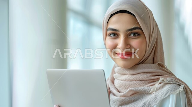 مفهوم الاهتمام بالمظهر الخارجي, صورة مقربة لامرأة عربية خليجية سعودية  محجبة مبتسمة تحمل بيدها جهاز الحاسوب المحمول, النظر الى الكاميرا بتعابير السرور, استخدام تقنيات الحديثة المتطورة.