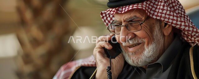 Using modern technology devices, talking to friends via social networking programs, chatting and conversations over the Internet, a close-up image of an elderly Saudi Arabian Gulf man wearing a traditional shemagh, thobe and glasses using a mobile phone, making voice calls with gestures of pleasure