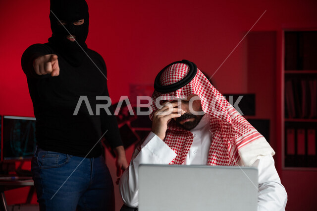 رجل عربي خليجي سعودي يجلس امام الحاسوب المحمول ، ايماءات الوجه واليد تدل على الخسارة والنصب، رجل ملثم يشير الى الكاميرا ، التعرض لسرقة بيانات مهمة ، هجوم الانترنت ، جرائم الكترونية ، الامن السيبراني