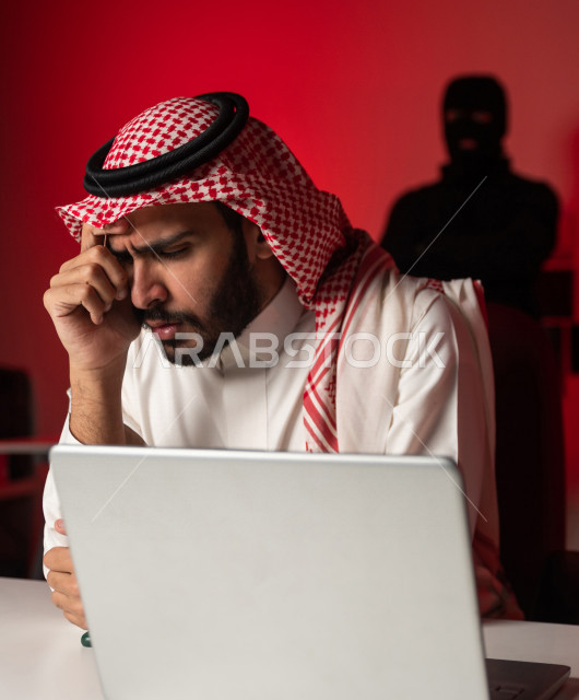 رجل عربي خليجي سعودي يجلس امام الحاسوب المحمول ، رجال ملثمين يقفون بالخلف ،رجل سعودي تعرض للنصب والخسارة، التعرض لهجمات سيبرانية، سرقة بيانات مهمة ،الجرائم الالكترونية، الامن السيبراني