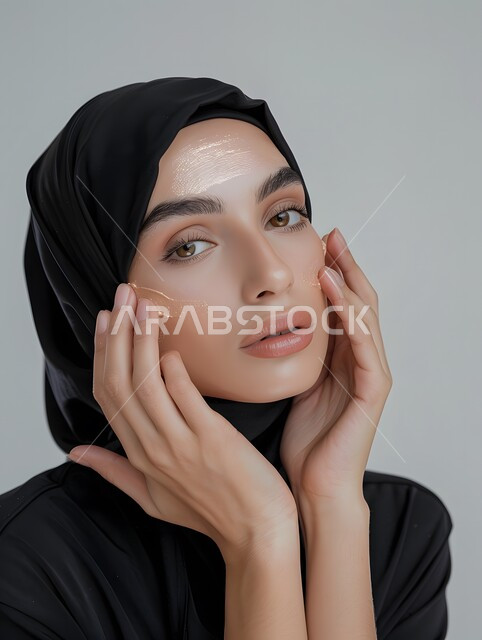 روتين وجلسات العناية بالبشرة للنساء, النظر الى الكاميرا بإيماءات السرور والبشاشة، مفهوم الجمال والاهتمام بالمظهر الخارجي,  بورتريه مقرب لامرأة عربية خليجية سعودية محجبة مبتسمة  تضع كريم الترطيب والتفتيح والتقشير, المحافظة على النضارة والشباب, خلفية رمادية