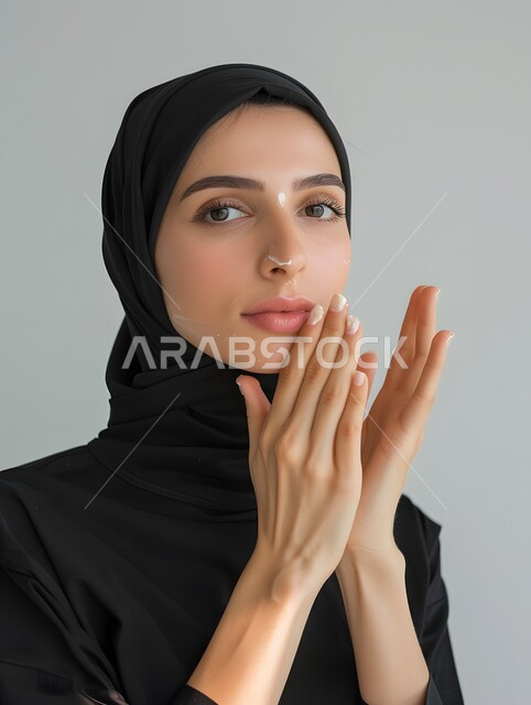 روتين وجلسات العناية بالبشرة للنساء, النظر الى الكاميرا بإيماءات السرور والبشاشة، مفهوم الجمال والاهتمام بالمظهر الخارجي,  بورتريه مقرب لامرأة عربية خليجية سعودية محجبة مبتسمة  تضع كريم الترطيب والتفتيح والتقشير, المحافظة على النضارة والشباب, خلفية رمادية