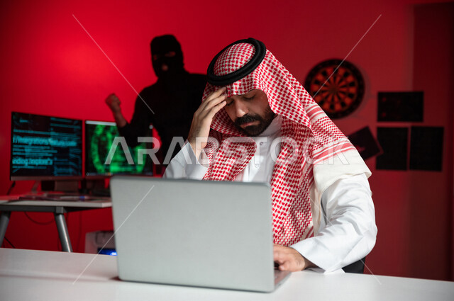 رجل عربي خليجي سعودي يجلس امام الحاسوب المحمول ، رجل رجل ملثم يقف بالخلف بايماءات تدل على النجاح والانتصار،رجل سعودي تعرض للنصب والاحتيال، التعرض لهجمات سيبرانية، سرقة بيانات مهمة ،الجرائم الالكترونية، الامن السيبراني