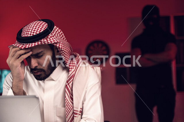رجل عربي خليجي سعودي يجلس امام الحاسوب المحمول ، رجال هكر يقفون بالخلف ،رجل سعودي محبط وحزين، التعرض لهجمات سيبرانية، سرقة بيانات مهمة ،الجرائم الالكترونية ، الامن السيبراني
