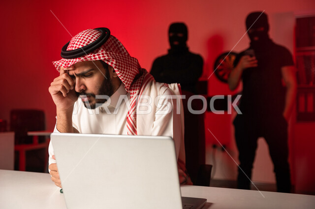 رجل عربي خليجي سعودي يجلس امام الحاسوب المحمول ، رجال ملثمين يقفون بالخلف ،رجل سعودي تعرض للنصب والاحتيال، التعرض لهجمات سيبرانية، سرقة بيانات مهمة ،الجرائم الالكترونية، الامن السيبراني
