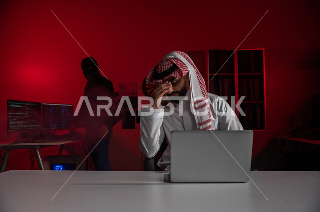 رجل عربي خليجي سعودي يجلس امام الحاسوب المحمول ، رجال هكر يقفون بالخلف ،رجل سعودي تعرض للخسارة والاحتيال، التعرض لهجمات سيبرانية، سرقة بيانات مهمة ،الجرائم الالكترونية ، الامن السيبراني