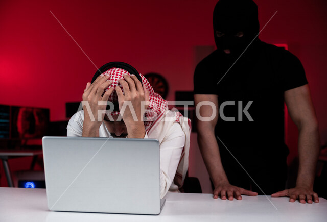رجل عربي خليجي سعودي يجلس امام الحاسوب المحمول ، رجال ملثمين يقفون بالخلف ،رجل سعودي تعرض للخسارة والنصب ، التعرض لهجوم الكتروني ، سرقة بيانات شخصية ،الجرائم الالكترونية، الامن السيبراني