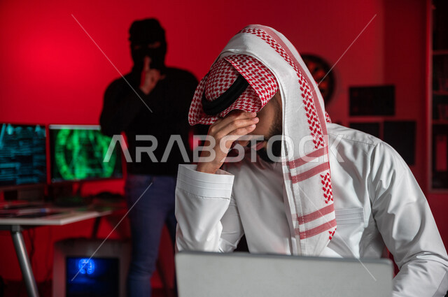 رجل عربي خليجي سعودي يجلس امام الحاسوب المحمول ، رجال ملثمين يقفون بالخلف ،رجل سعودي تعرض للنصب والخسارة، التعرض لهجمات سيبرانية، سرقة بيانات مهمة ،الجرائم الالكترونية ، الامن السيبراني