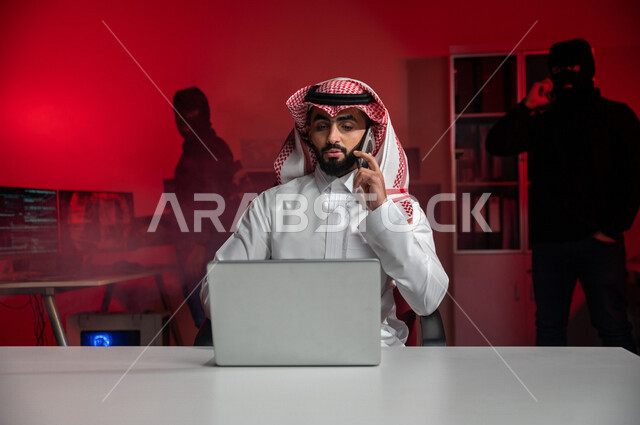 رجل عربي خليجي سعودي يجلس امام الحاسوب المحمول ، يتحدث عبر الهاتف المحمول ،  رجل ملثم يستخدم الهاتف المحمول ،التنصت على المكالمة ،التسلل عبر الشبكات الهاتفية ، جرائم الكترونية ، ابتزاز وتهديد، الامن السيبراني