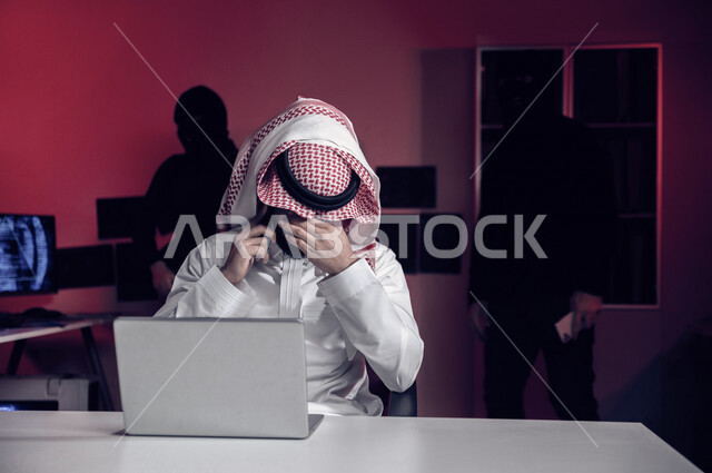 رجل عربي خليجي سعودي يجلس امام الحاسوب المحمول ، رجال هكر يقفون بالخلف ،رجل سعودي تعرض للخسارة والنصب، التعرض لهجمات سيبرانية، سرقة بيانات مهمة ،الجرائم الالكترونية، الامن السيبراني