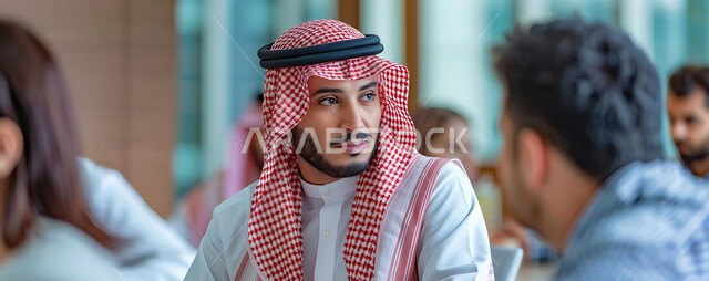 مفهوم الأناقة والاهتمام بالمظهر الخارجي، صورة مقربة لشاب عربي خليجي سعودي يرتدي الشماغ والثوب التقليدي يجلس مع أصدقائه على طاولة ويحاول النظر الى شيء ما، الاستمتاع بقضاء الأوقات الممتعة مع الأصدقاء