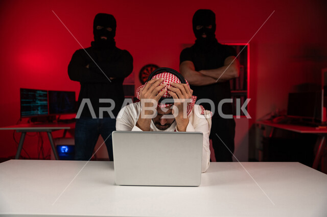 A Saudi Arabian Gulf man sitting in front of a laptop, masked men standing behind, a Saudi man exposed to loss and fraud, exposure to electronic attack, personal data theft, electronic crimes, cyber security